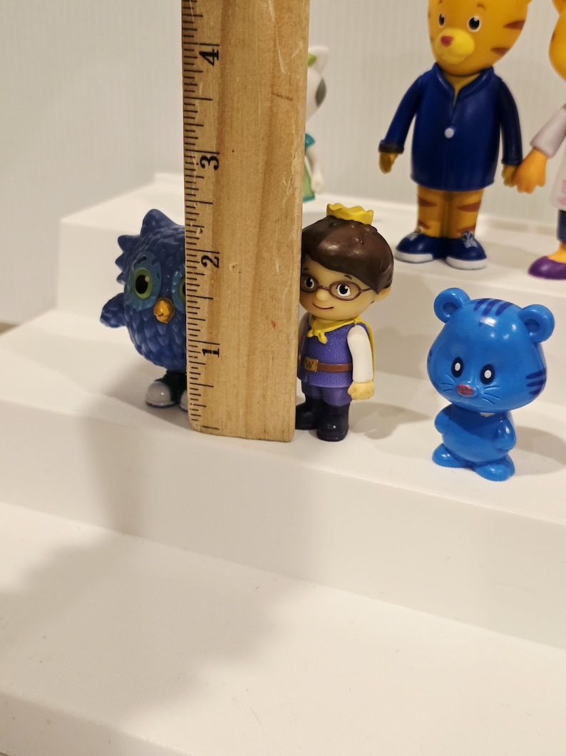 May include: A collection of small plastic character figurines including a blue owl, a figure with a gold crown, and a blue bear, positioned next to a wooden ruler showing a height of approximately 4 inches for the tallest visible figure.