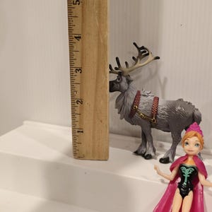May include: A wooden ruler next to a grey reindeer figurine with brown antlers and a red harness. A small doll of a character wearing a pink cape and a black and green outfit is also present.