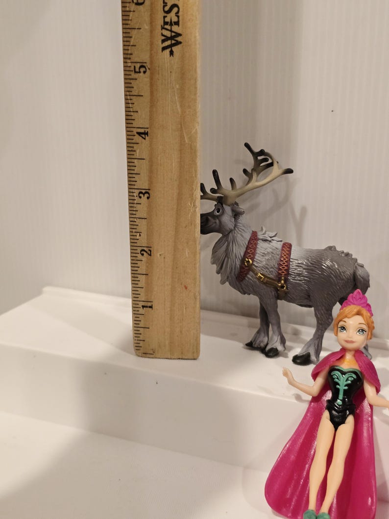 May include: A wooden ruler next to a grey reindeer figurine with brown antlers and a red harness. A small doll of a character wearing a pink cape and a black and green outfit is also present.