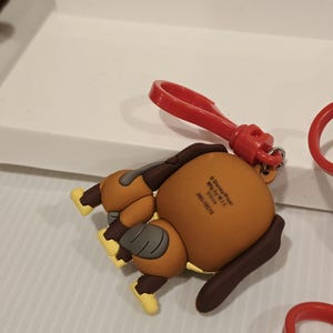 May include: A brown and tan cartoon dog keyring with a red clip. The dog is lying down with its rear end facing the viewer. The keyring has yellow accents on its feet and a brown ear. The clip is attached to the dog's head.
