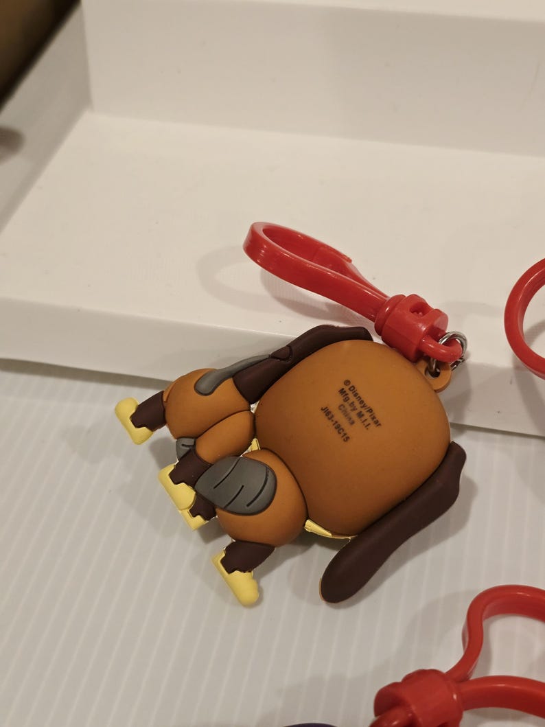 May include: A brown and tan cartoon dog keyring with a red clip. The dog is lying down with its rear end facing the viewer. The keyring has yellow accents on its feet and a brown ear. The clip is attached to the dog's head.