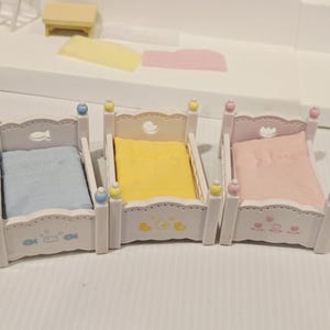 Puede incluir: Tres camas blancas en miniatura con ropa de cama azul pastel, amarilla y rosa. Cada cama tiene detalles decorativos, incluyendo peces, patos y corazones. Las camas son parte de un juego de casa de mu&ntilde;ecas.