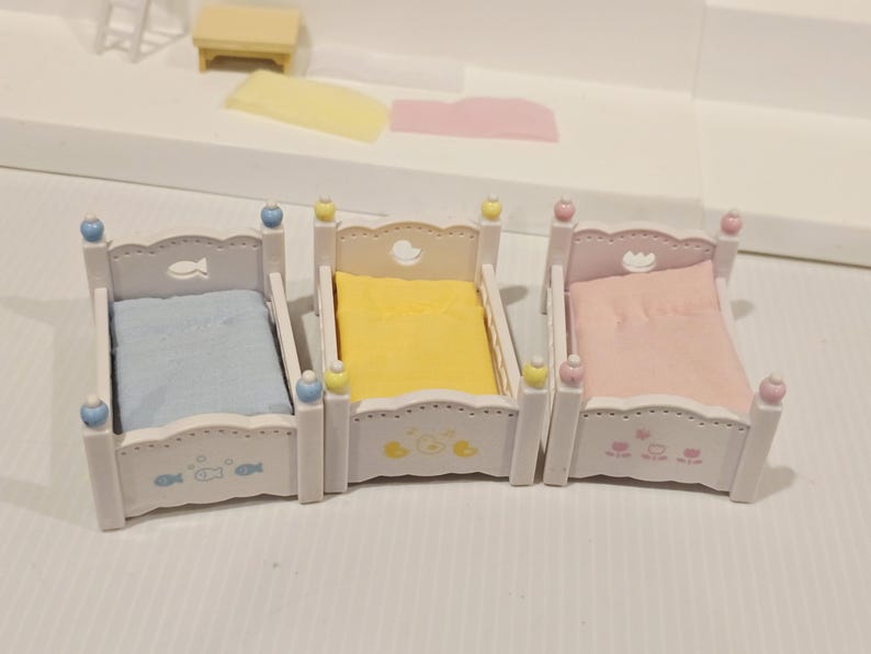 Puede incluir: Tres camas blancas en miniatura con ropa de cama azul pastel, amarilla y rosa. Cada cama tiene detalles decorativos, incluyendo peces, patos y corazones. Las camas son parte de un juego de casa de mu&ntilde;ecas.