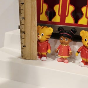 K&ouml;nnte beinhalten: Ein Holzlineal neben drei kleinen Figuren von Zeichentrickfiguren. Die Figuren tragen rote Jacken und Kleider. Das Lineal misst etwa 7,6 cm in der H&ouml;he.