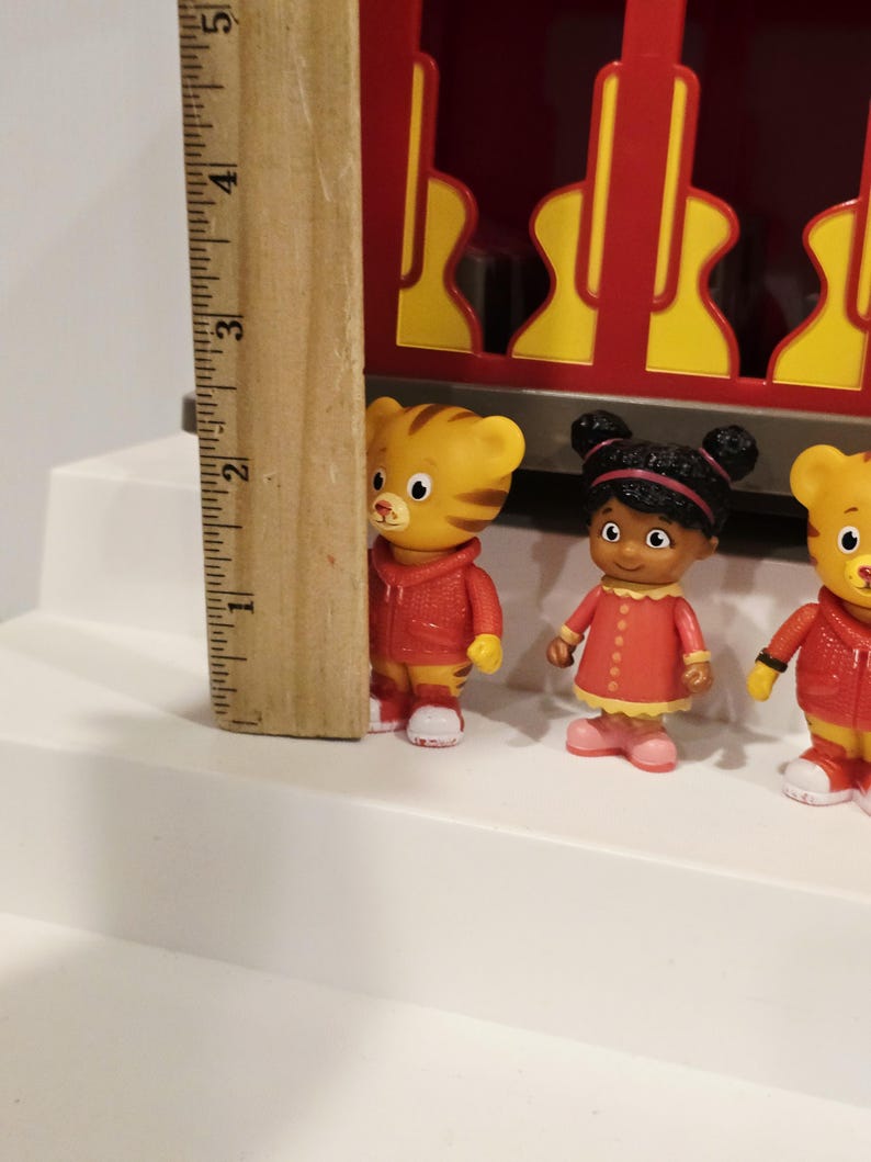 K&ouml;nnte beinhalten: Ein Holzlineal neben drei kleinen Figuren von Zeichentrickfiguren. Die Figuren tragen rote Jacken und Kleider. Das Lineal misst etwa 7,6 cm in der H&ouml;he.