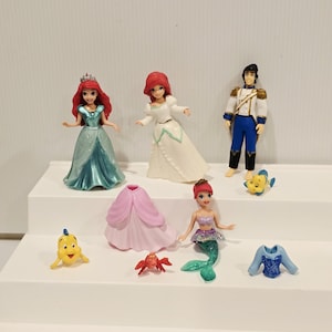 K&ouml;nnte beinhalten: Eine Sammlung von Disney's Die kleine Meerjungfrau Figuren. Das Set enth&auml;lt Arielle in einem t&uuml;rkisfarbenen Kleid, Arielle in einem wei&szlig;en Kleid, Prinz Eric, Fabius, ein rosa Kleid, Sebastian, eine Meerjungfrau Arielle und ein blaues Oberteil.