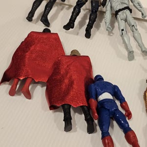 May include: A collection of action figures featuring various superhero characters. The figures are in different poses and colors, including black, blue, red, and gray. Some figures have capes, and others have detailed costumes.