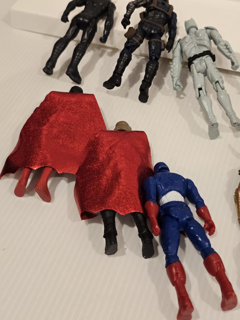 May include: A collection of action figures featuring various superhero characters. The figures are in different poses and colors, including black, blue, red, and gray. Some figures have capes, and others have detailed costumes.