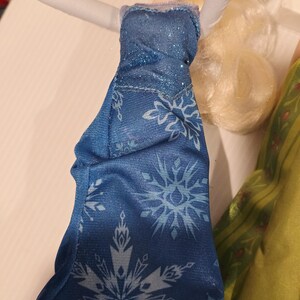 May include: A doll in a blue dress featuring a snowflake design. The dress has a sparkly bodice and a flowing skirt. The doll has blonde hair and is posed with arms raised.