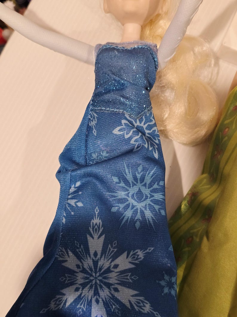 May include: A doll in a blue dress featuring a snowflake design. The dress has a sparkly bodice and a flowing skirt. The doll has blonde hair and is posed with arms raised.