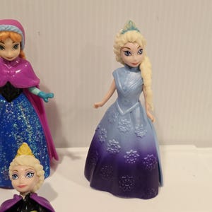 May include: A collection of small plastic figurines of characters from the animated film "Frozen". The figurines include Elsa, Anna, and other characters. Elsa is wearing a blue dress with a purple ombre skirt and a tiara.