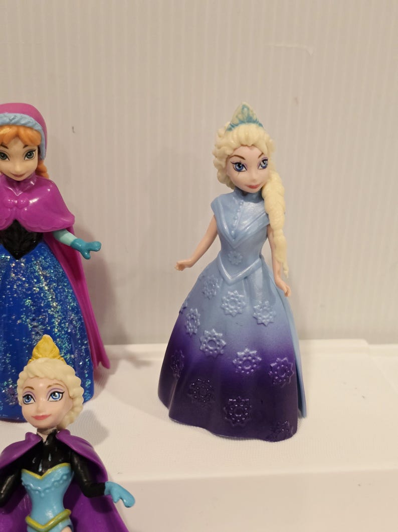May include: A collection of small plastic figurines of characters from the animated film "Frozen". The figurines include Elsa, Anna, and other characters. Elsa is wearing a blue dress with a purple ombre skirt and a tiara.