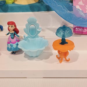 May include: Ariel's Secret Play Set featuring a miniature Ariel doll with red hair and a blue and pink dress. The set includes a light blue sink, an orange table with a blue lamp, and other decorative elements. The packaging is visible in the background.