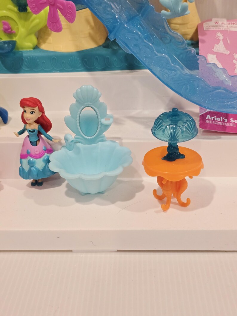 May include: Ariel's Secret Play Set featuring a miniature Ariel doll with red hair and a blue and pink dress. The set includes a light blue sink, an orange table with a blue lamp, and other decorative elements. The packaging is visible in the background.