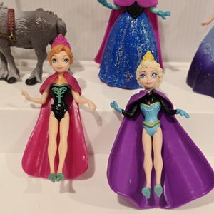 May include: A collection of small plastic dolls, featuring characters from an animated film. Two dolls are in the foreground, one with a pink cape and black bodysuit, the other with a purple cape and blue bodysuit. Other dolls are visible.