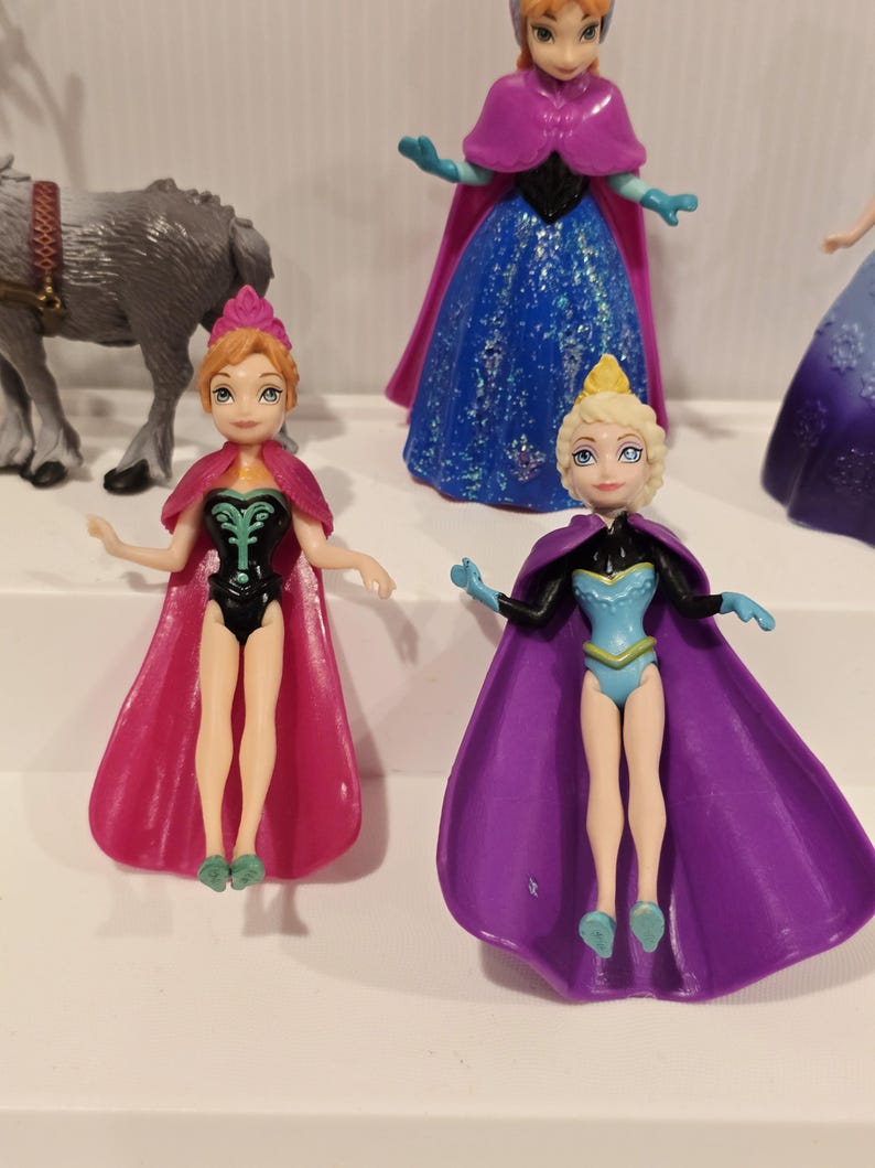 May include: A collection of small plastic dolls, featuring characters from an animated film. Two dolls are in the foreground, one with a pink cape and black bodysuit, the other with a purple cape and blue bodysuit. Other dolls are visible.