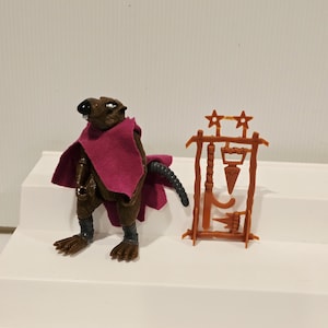 May include: A brown anthropomorphic rat action figure wearing a magenta fabric cape, standing next to a brown plastic accessory rack containing various small molded weapons and two star-shaped projectiles at the top of the frame.