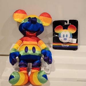 May include: A rainbow-colored Mickey Mouse plush toy and a matching keychain. The plush toy features a vibrant spectrum of colors across its body, including red, orange, yellow, green, blue, and purple. The keychain has a Mickey Mouse face with rainbow ears. The Disney Collection logo is visible.