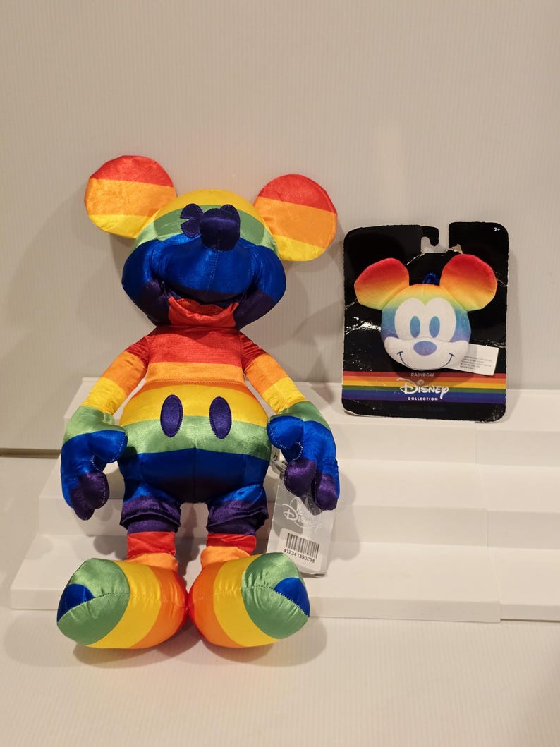 May include: A rainbow-colored Mickey Mouse plush toy and a matching keychain. The plush toy features a vibrant spectrum of colors across its body, including red, orange, yellow, green, blue, and purple. The keychain has a Mickey Mouse face with rainbow ears. The Disney Collection logo is visible.