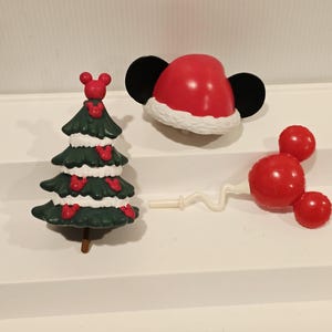 May include: A dark green Christmas tree figurine with white snow accents and small red Mickey Mouse head icons, a red Santa hat with black mouse ears, and a red Mickey Mouse head balloon on a white wavy stick, all made of smooth plastic material.