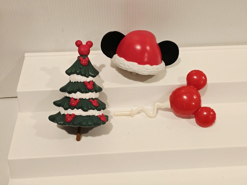 May include: A dark green Christmas tree figurine with white snow accents and small red Mickey Mouse head icons, a red Santa hat with black mouse ears, and a red Mickey Mouse head balloon on a white wavy stick, all made of smooth plastic material.