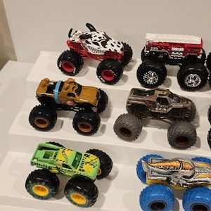 Op de afbeelding: Een verzameling speelgoed monstertrucks in verschillende kleuren en ontwerpen. De trucks hebben oversized banden en gedetailleerde graphics, waaronder een truck met Dalmatische thema, een Scooby-Doo truck en een groene truck met het woord "BURSTER".