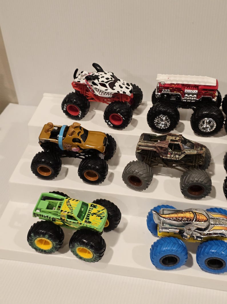 Op de afbeelding: Een verzameling speelgoed monstertrucks in verschillende kleuren en ontwerpen. De trucks hebben oversized banden en gedetailleerde graphics, waaronder een truck met Dalmatische thema, een Scooby-Doo truck en een groene truck met het woord "BURSTER".