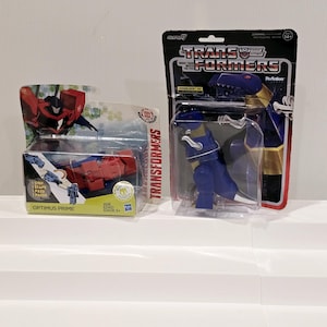 May include: Two Transformers action figures in their packaging. The figure on the left is Optimus Prime, a red robot with a blue truck. The figure on the right is a blue and gold robot with a dinosaur head.