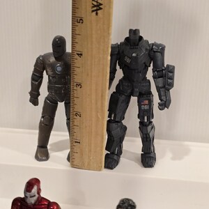 May include: Two Iron Man action figures, one bronze and one black, stand next to a wooden ruler. The black figure is approximately 5 inches tall. Other figures are visible in the foreground.