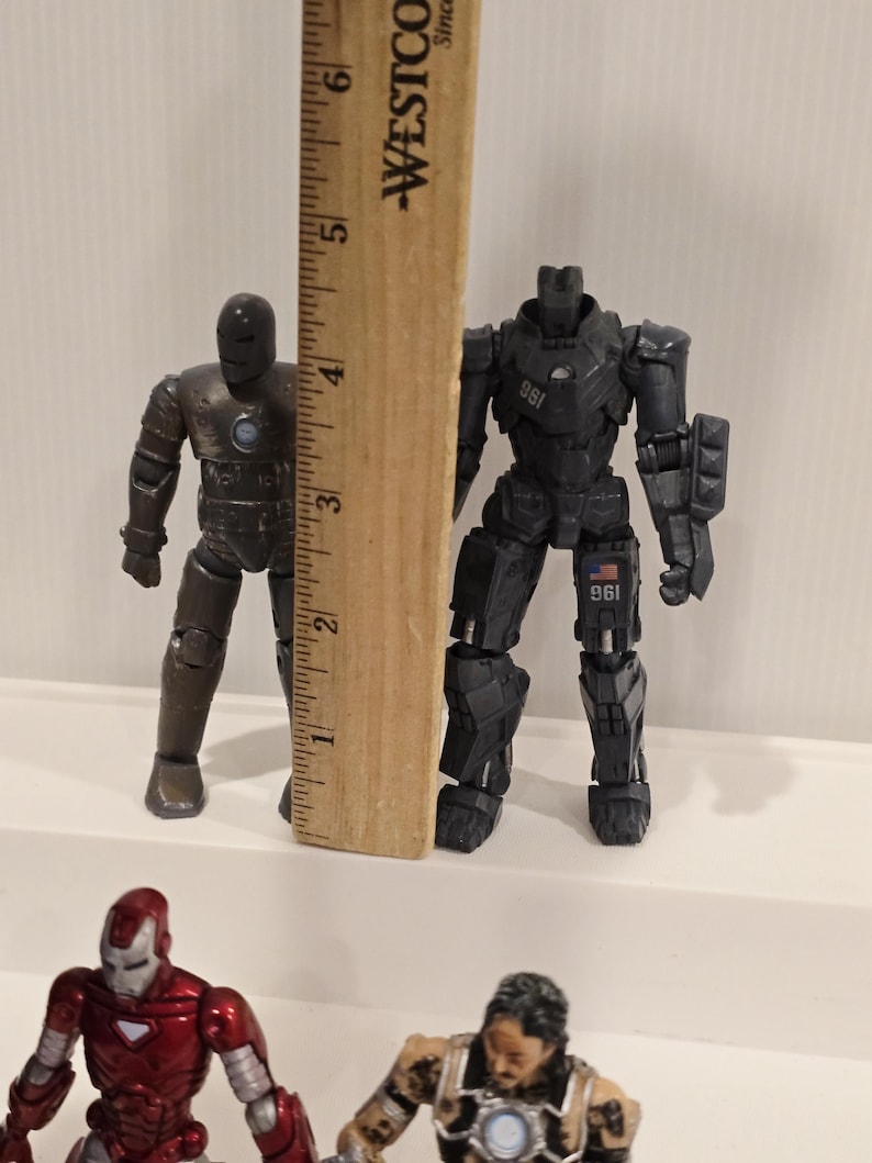 May include: Two Iron Man action figures, one bronze and one black, stand next to a wooden ruler. The black figure is approximately 5 inches tall. Other figures are visible in the foreground.