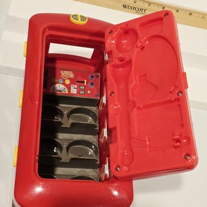 K&ouml;nnte beinhalten: Ein roter Spielzeugbus mit einer offenen Seite, die ein Fach mit Formen und einem Bedienfeld freilegt. Die offene T&uuml;r hat geformte Formen. Gelbe und silberne Akzente sind sichtbar. Das Spielzeug ist f&uuml;r Kinder konzipiert.