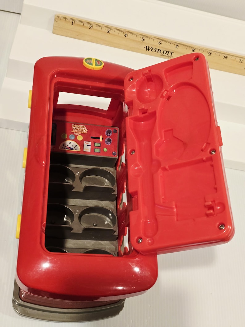 K&ouml;nnte beinhalten: Ein roter Spielzeugbus mit einer offenen Seite, die ein Fach mit Formen und einem Bedienfeld freilegt. Die offene T&uuml;r hat geformte Formen. Gelbe und silberne Akzente sind sichtbar. Das Spielzeug ist f&uuml;r Kinder konzipiert.