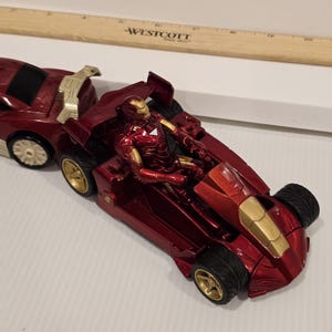 May include: A red and gold Iron Man action figure in a red race car. The car has gold wheels and accents. The figure is in a seated position inside the car. The car is connected to a red trailer.