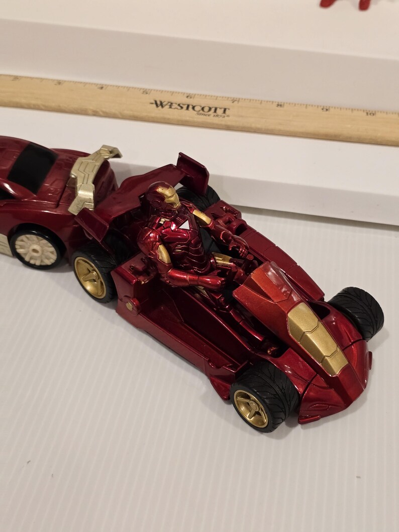 May include: A red and gold Iron Man action figure in a red race car. The car has gold wheels and accents. The figure is in a seated position inside the car. The car is connected to a red trailer.