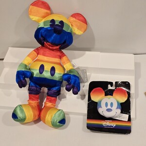 May include: A rainbow-colored Mickey Mouse plush toy and a matching face-shaped plush on a black backing. The plush toy has rainbow ears, a blue face, and a rainbow-striped body. The backing has the words "Rainbow Collection" and the Disney logo.
