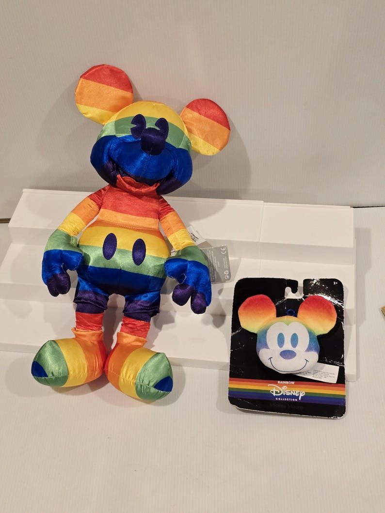 May include: A rainbow-colored Mickey Mouse plush toy and a matching face-shaped plush on a black backing. The plush toy has rainbow ears, a blue face, and a rainbow-striped body. The backing has the words "Rainbow Collection" and the Disney logo.