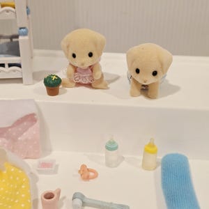 Puede incluir: Dos peque&ntilde;as figuras de cachorros color canela con ojos negros, una con un vestido rosa y la otra con un pa&ntilde;al azul. Tambi&eacute;n hay biberones, un chupete y otros accesorios de beb&eacute; en miniatura.