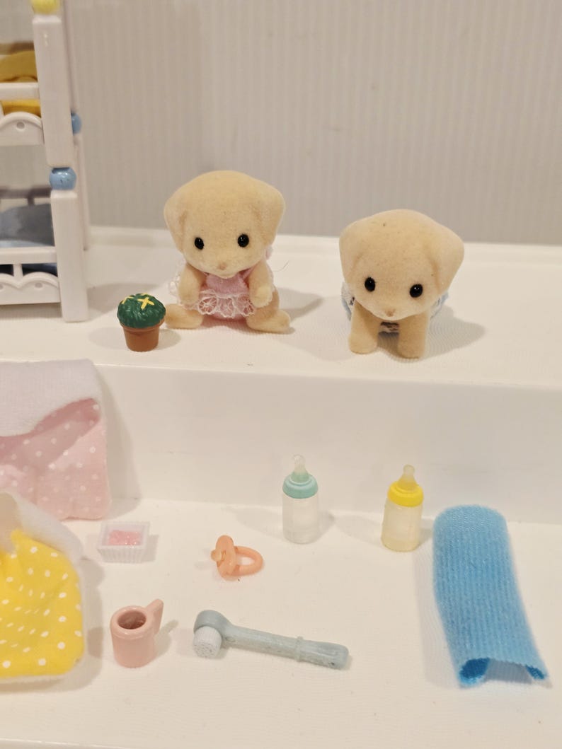 Puede incluir: Dos peque&ntilde;as figuras de cachorros color canela con ojos negros, una con un vestido rosa y la otra con un pa&ntilde;al azul. Tambi&eacute;n hay biberones, un chupete y otros accesorios de beb&eacute; en miniatura.
