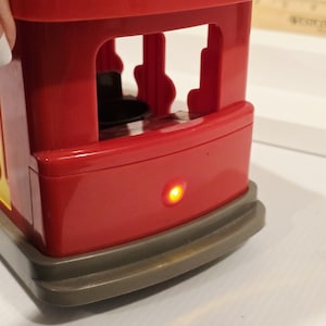 K&ouml;nnte beinhalten: Eine rote Spielzeugstra&szlig;enbahn mit gelbem Akzent und grauem Sockel. Die Stra&szlig;enbahn hat ein offenes Design mit einer kleinen, runden &Ouml;ffnung im Inneren. Ein kleines, gelbes Licht leuchtet an der Vorderseite der Stra&szlig;enbahn.