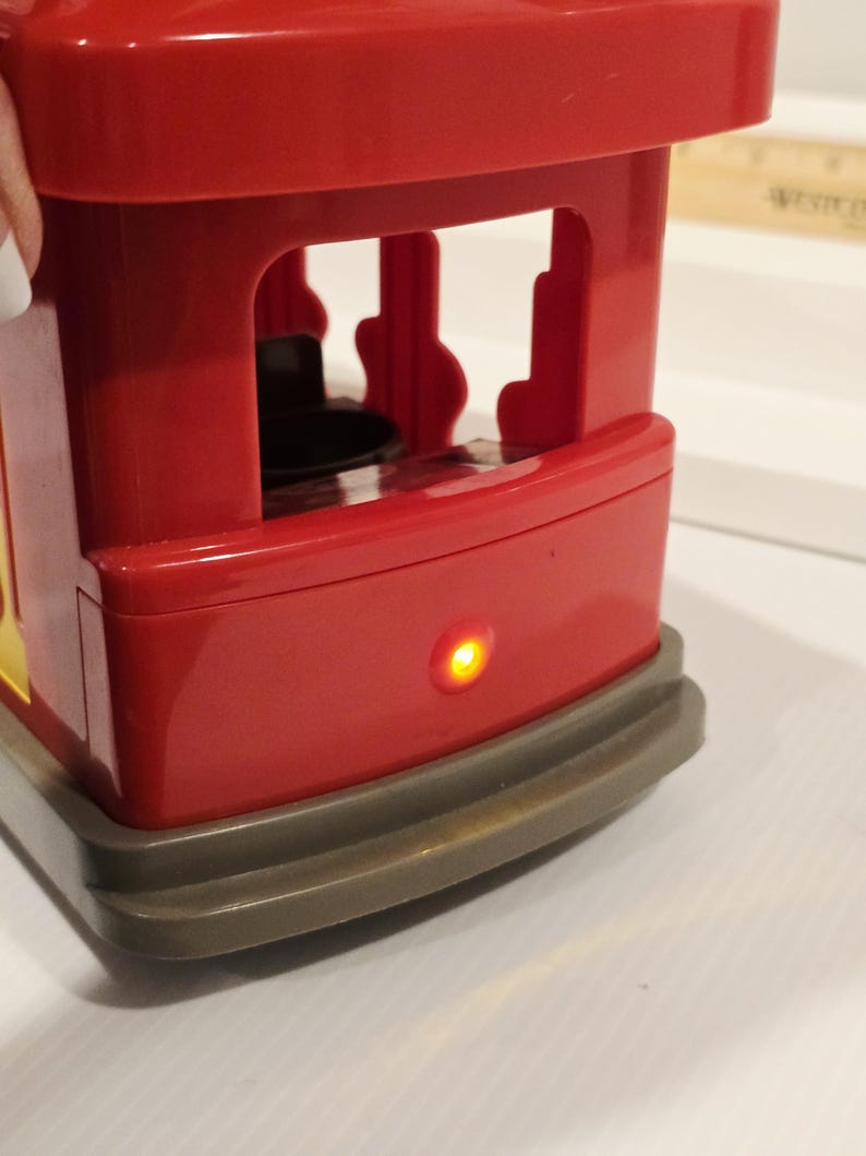 K&ouml;nnte beinhalten: Eine rote Spielzeugstra&szlig;enbahn mit gelbem Akzent und grauem Sockel. Die Stra&szlig;enbahn hat ein offenes Design mit einer kleinen, runden &Ouml;ffnung im Inneren. Ein kleines, gelbes Licht leuchtet an der Vorderseite der Stra&szlig;enbahn.
