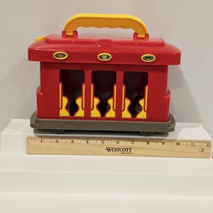 K&ouml;nnte beinhalten: Ein roter Spielzeugzug mit gelbem Griff und R&auml;dern. Der Zug hat drei &Ouml;ffnungen mit gelben gitarrenf&ouml;rmigen Ausschnitten. Der Zug ist etwa 30 cm lang.