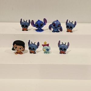 May include: A collection of eight small figurines featuring characters from the animated film "Lilo & Stitch." The figurines are primarily blue, with some having orange accents. One figurine depicts a girl with black hair and a brown dress.