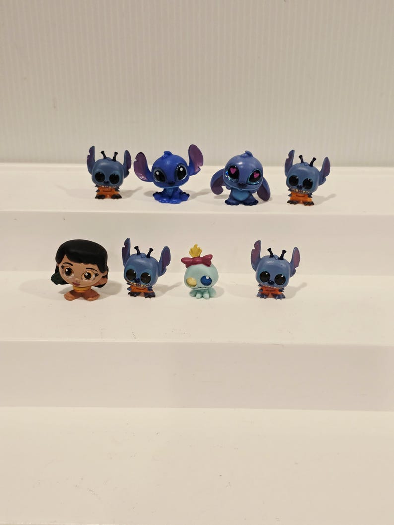 May include: A collection of eight small figurines featuring characters from the animated film "Lilo & Stitch." The figurines are primarily blue, with some having orange accents. One figurine depicts a girl with black hair and a brown dress.
