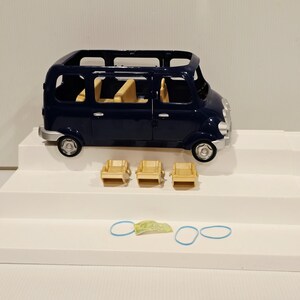May include: A navy blue toy van with a silver bumper and wheels. Three small tan chairs are in front of the van, along with two blue rubber bands and a small green and yellow object. The van has tan seats inside.