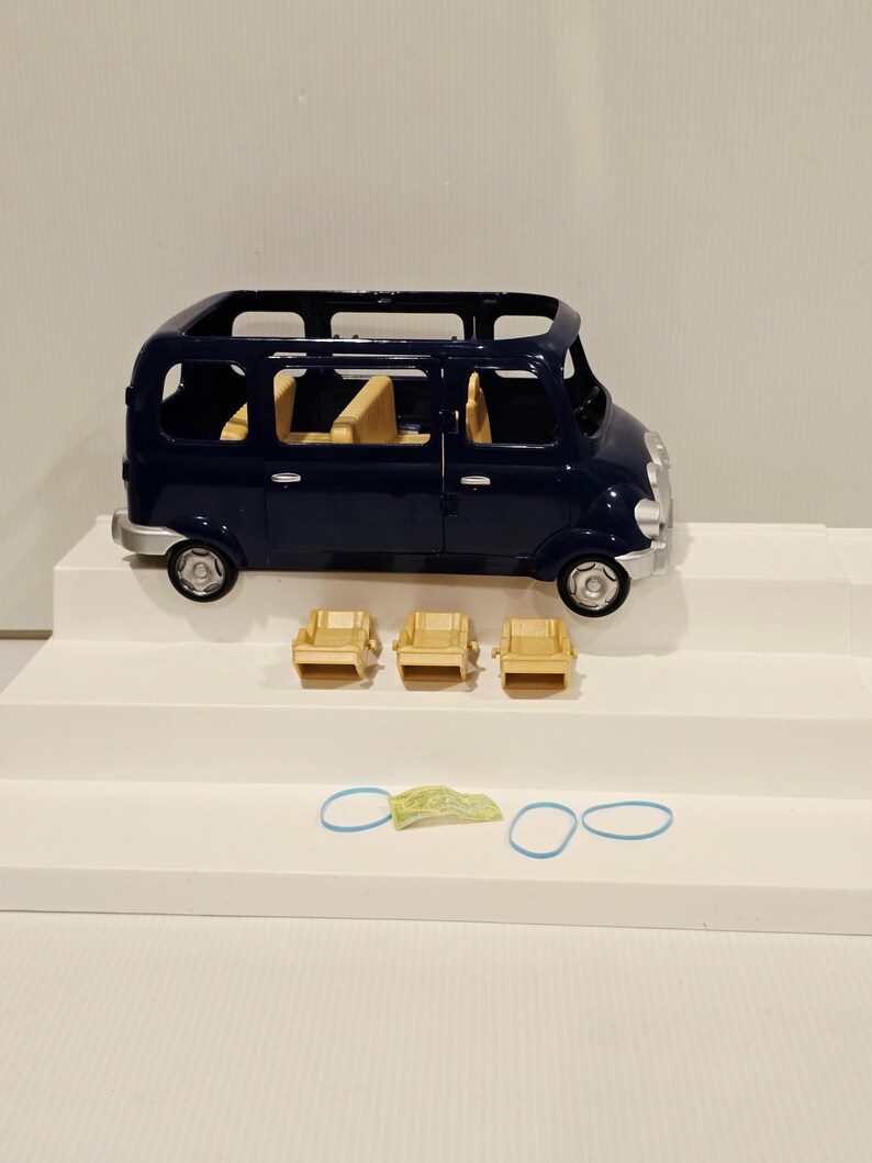 May include: A navy blue toy van with a silver bumper and wheels. Three small tan chairs are in front of the van, along with two blue rubber bands and a small green and yellow object. The van has tan seats inside.