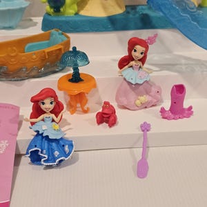 K&ouml;nnte beinhalten: Eine Sammlung farbenfroher Spielzeugfiguren und Zubeh&ouml;rteile aus dem Film Arielle, die Meerjungfrau. Enth&auml;lt Ariel-Puppen in blauen und rosa Kleidern, eine rote Krabbe, ein kleines orangefarbenes Boot, einen Tisch mit einer Lampe und andere thematische Teile.
