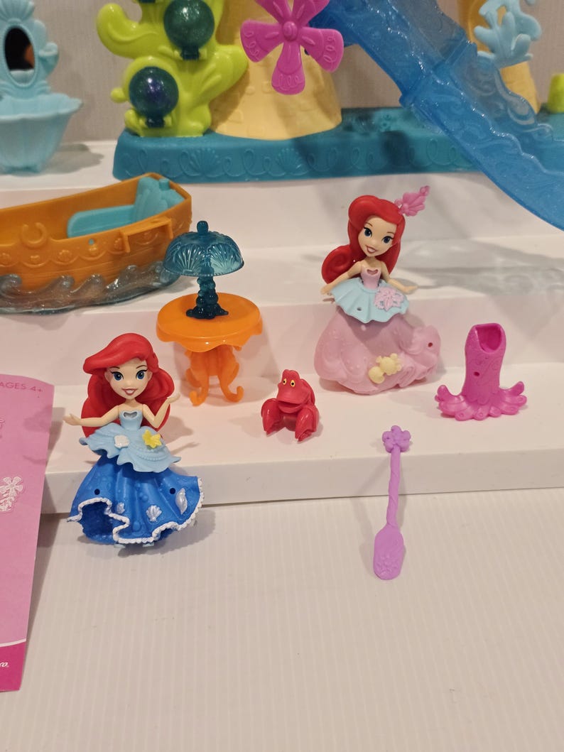 K&ouml;nnte beinhalten: Eine Sammlung farbenfroher Spielzeugfiguren und Zubeh&ouml;rteile aus dem Film Arielle, die Meerjungfrau. Enth&auml;lt Ariel-Puppen in blauen und rosa Kleidern, eine rote Krabbe, ein kleines orangefarbenes Boot, einen Tisch mit einer Lampe und andere thematische Teile.