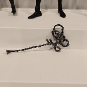 May include: A silver-colored, twisted metal staff with a decorative, looped top and a pointed end. The staff is lying on a white surface, with parts of action figures visible in the background.