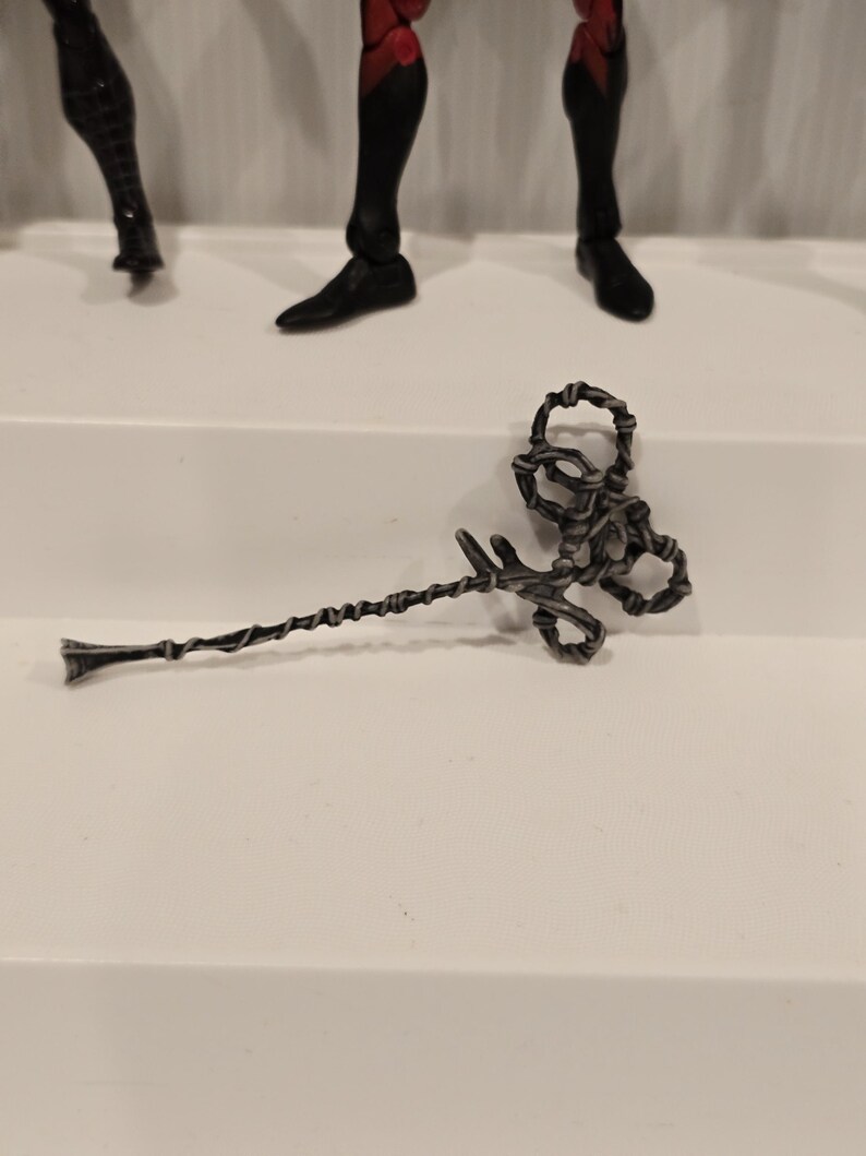 May include: A silver-colored, twisted metal staff with a decorative, looped top and a pointed end. The staff is lying on a white surface, with parts of action figures visible in the background.