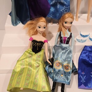 May include: Two Anna dolls from the film Frozen. One doll wears a black top with gold detailing and a green skirt with pink floral patterns. The other doll wears a blue dress with sunflower details. Other dolls and accessories are visible.